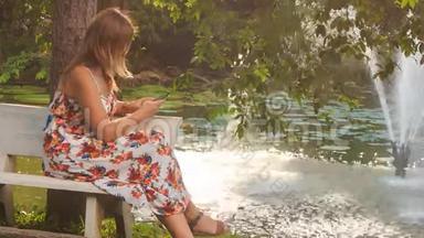 金发女郎坐在池塘的长凳<strong>上</strong>，在公园用手机拍照